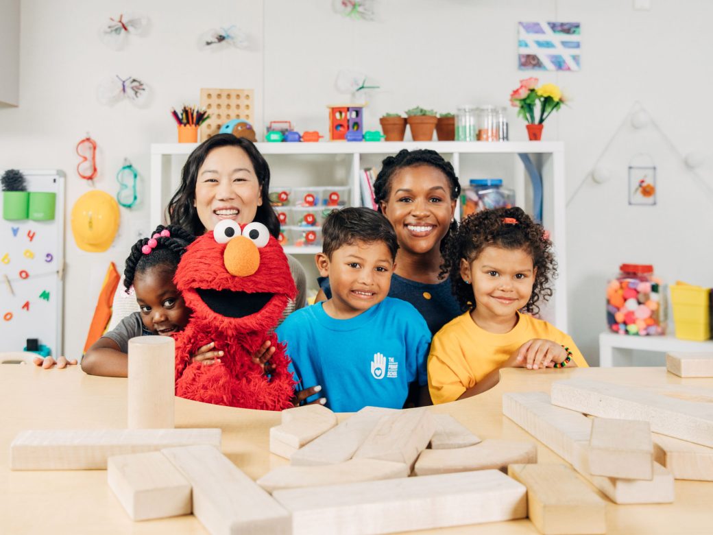 Elmo, Priscilla Chan, a teacher, and students from The Primary School sit at a table covered in building blocks.