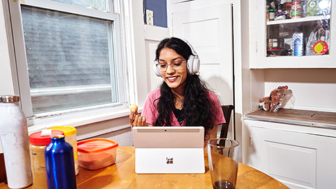 Sabiha in her dining room eating a timbit while using her surface tablet and wearing headphones