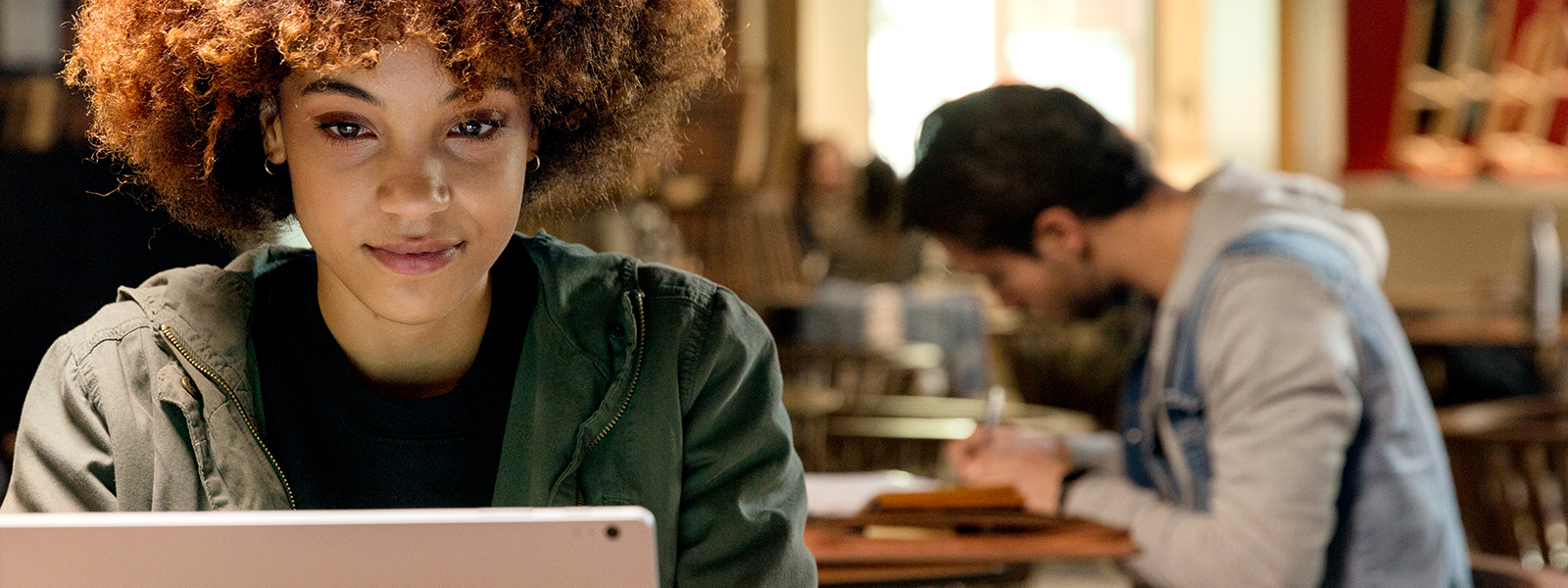 A woman studying at the computer with a man in the background taking notes.