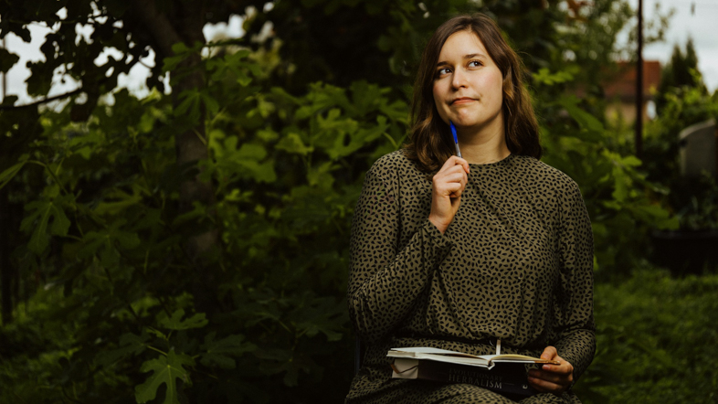 A woman sitting on a bench with a notebook in her lap, holding a blue pen