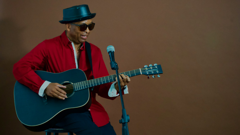 A man wearing a hat, sitting on a stool, playing a guitar