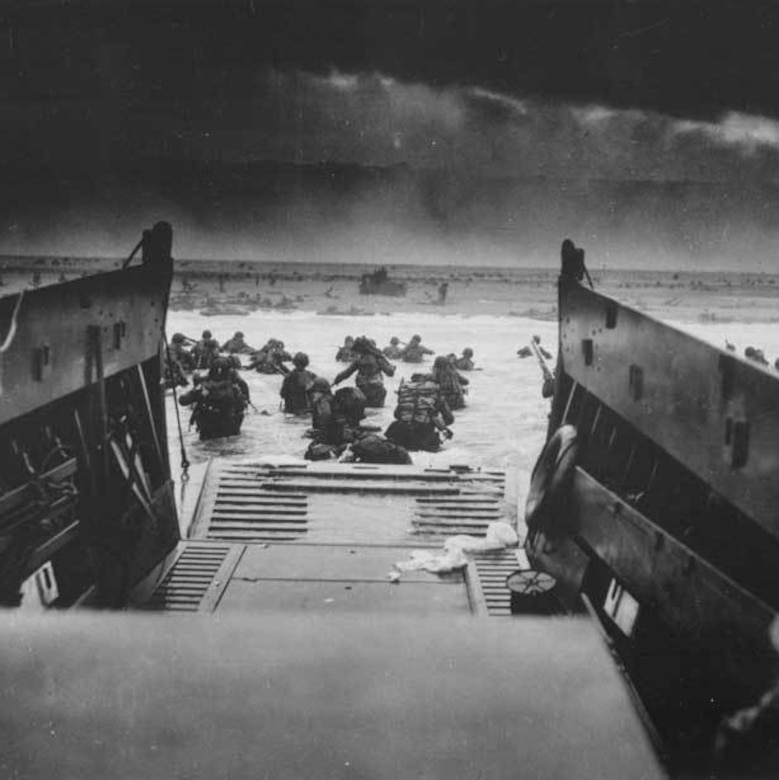 Soldiers wade through water while making their way from a landing craft ramp onto shore.