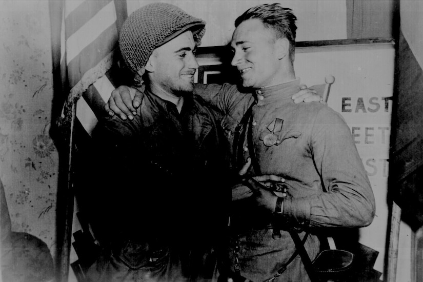 Two men in military uniforms smile at each other.