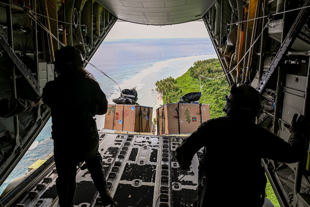 Airman watch as two packages with parachutes attached fall out the back of an aircraft during flight over coastline.