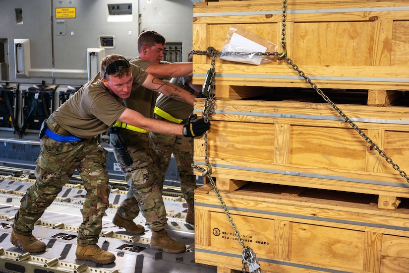 Three airmen push a pallet of weapons cargo.