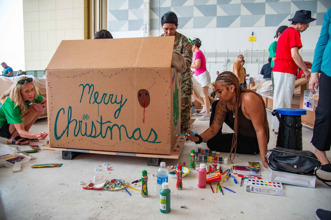 Service members and civilians decorate a large box with the words ‘Merry Christmas’ on its side. Other people stand and work around them in a brightly lit room.