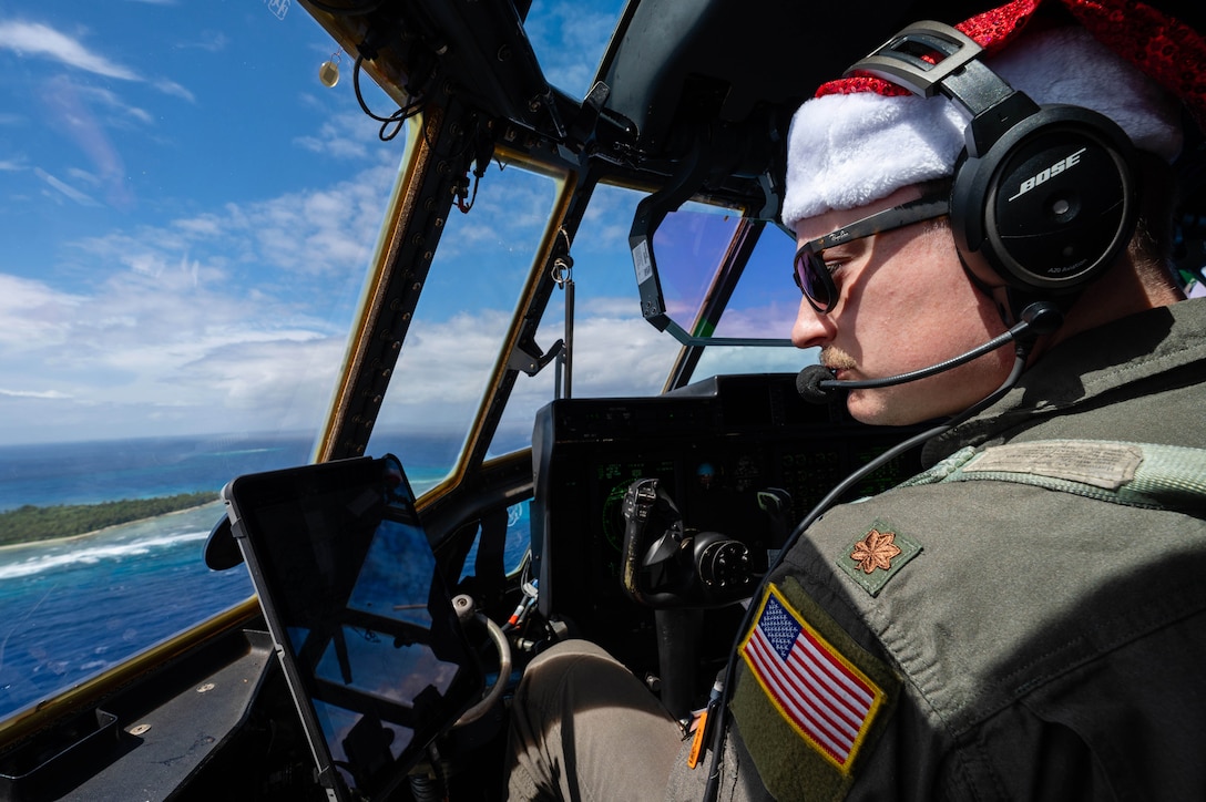 An irman is seen inside a plane wearing a Santa hat with blue skies and a small island and ocean below.