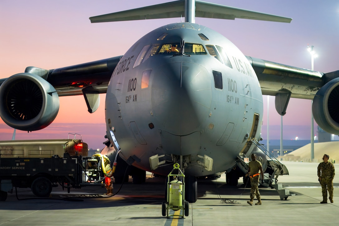 A close-up of the front of an aircraft parked on a tarmac as service members walk by under a pinkish sky.