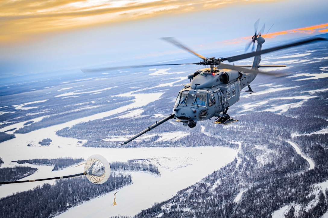 A military helicopter flies in a light blue, yellow and purple sky above snowy and icy wooded terrain during a refueling exercise.