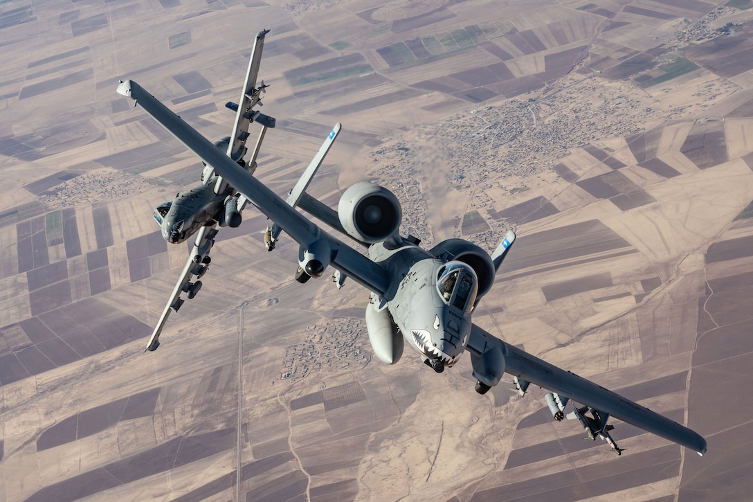 Two aircraft tilting in opposite directions fly over a desert-like area during daylight.