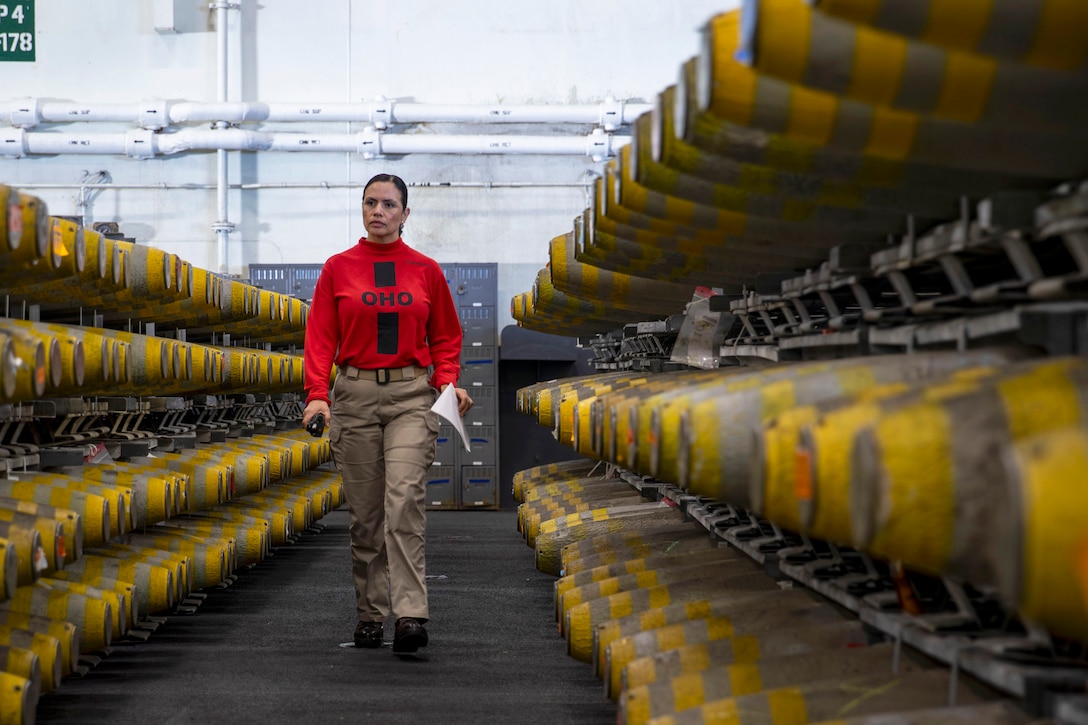 A sailor walks through a row of ammunition stacked on shelves in a warehouse aboard ship.