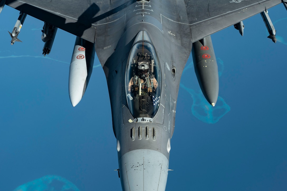 A pilot flies a jet over blue water, as seen from above.