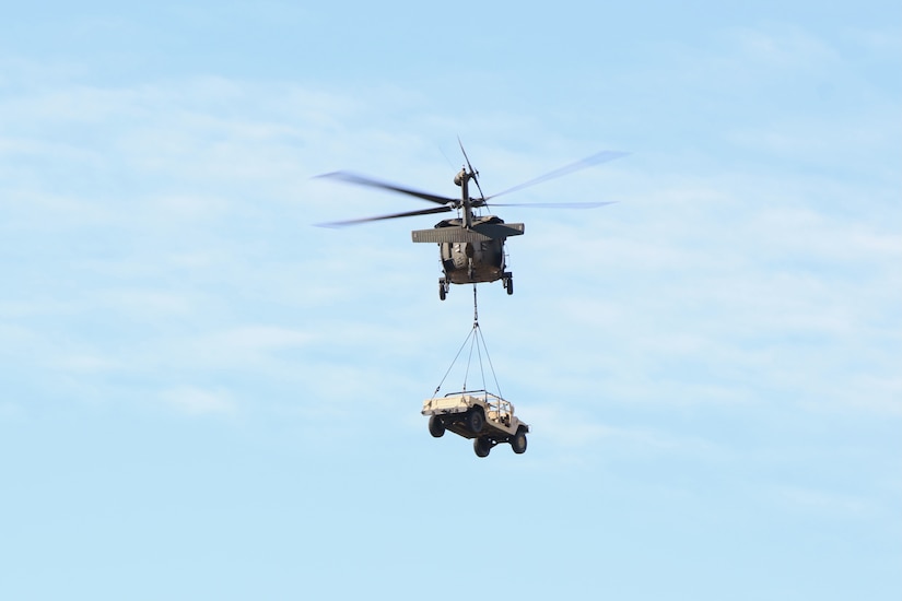 A helicopter with a military vehicle suspended from it by a line flies through blue sky.