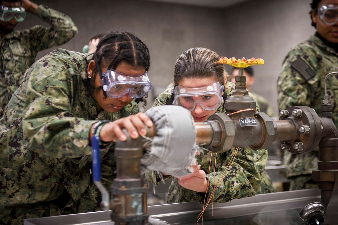 Sailors wearing protective eyewear work on a valve as others in a concrete-like structure.