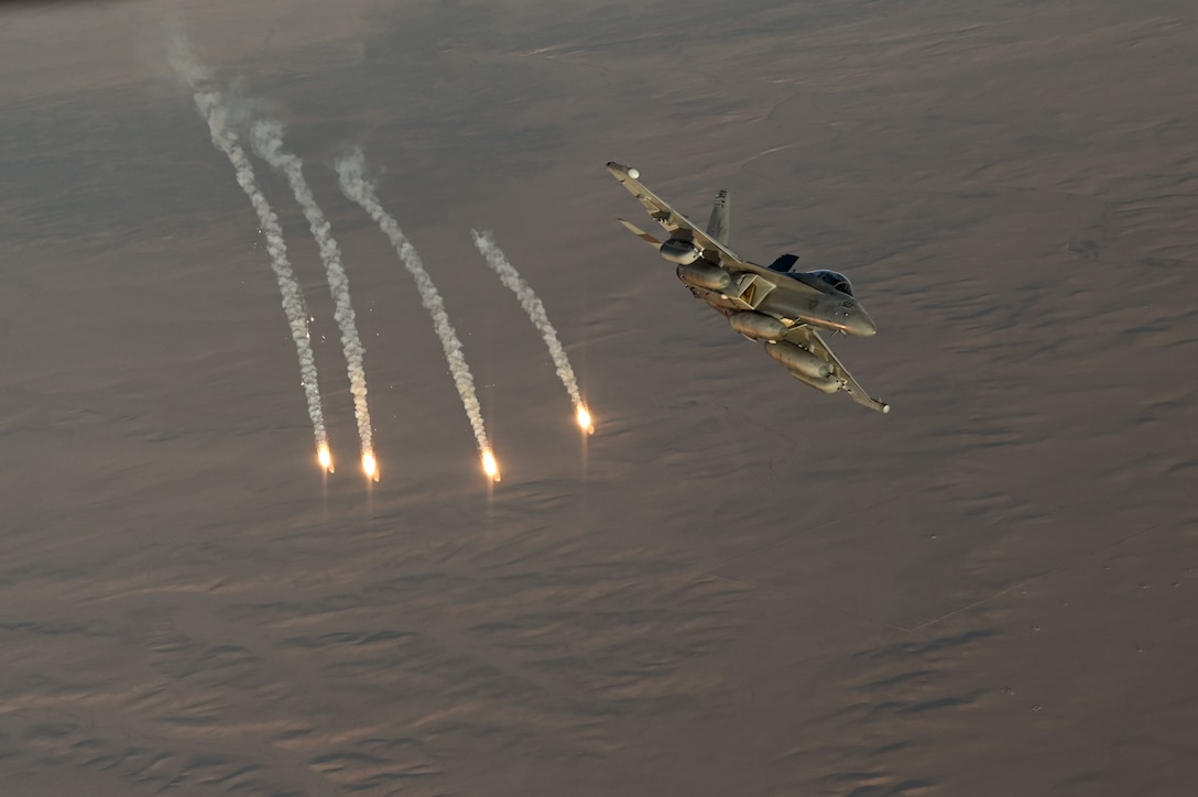 Four flares descend in the sky next to a jet in flight over desert-like terrain.