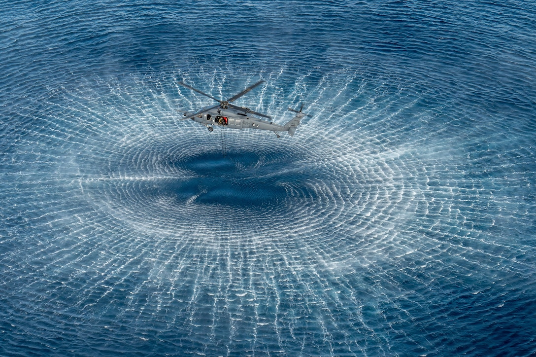 An airman holds a ladder out the open door of a helicopter hovering over blue water, creating a circular wake in the water.