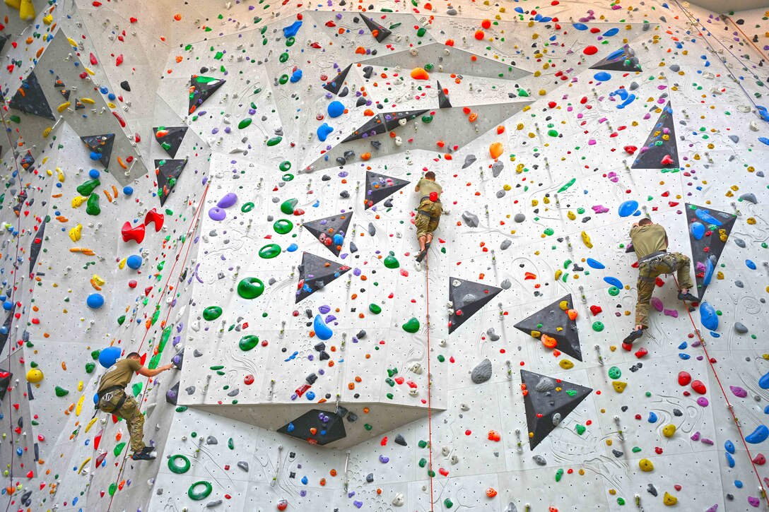 Three service members wearing harnesses climb a colorful indoor rock wall as seen from below.