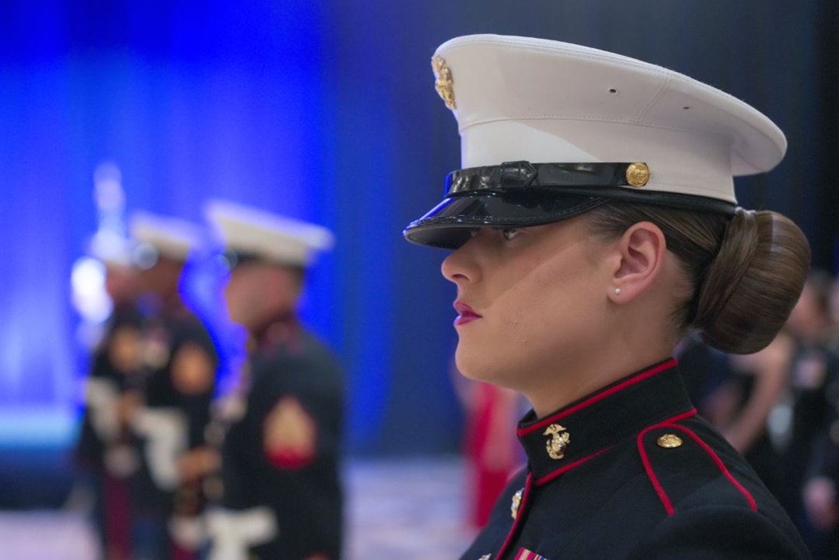 A close-up of a Marine’s face while standing at attention in a ceremonial uniform with blurred Marines in the background.