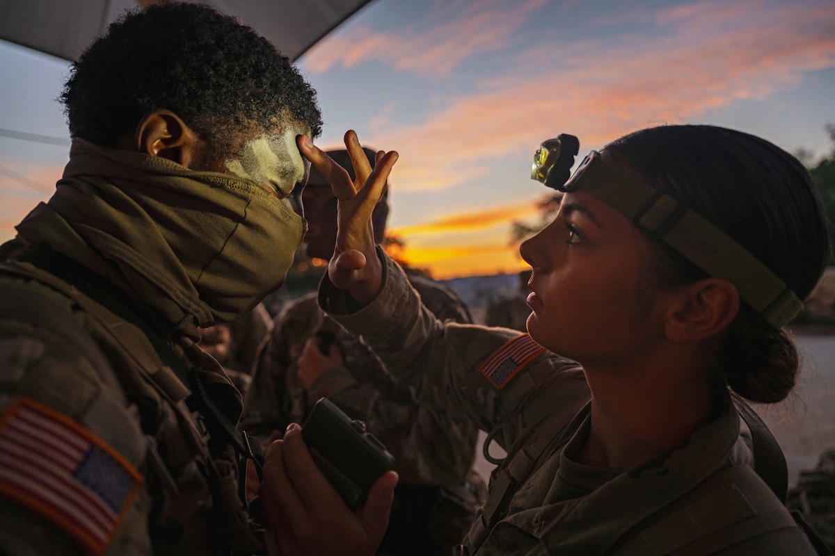 A soldier wearing a headlamp applies camouflage to another soldier under a sunlit sky.