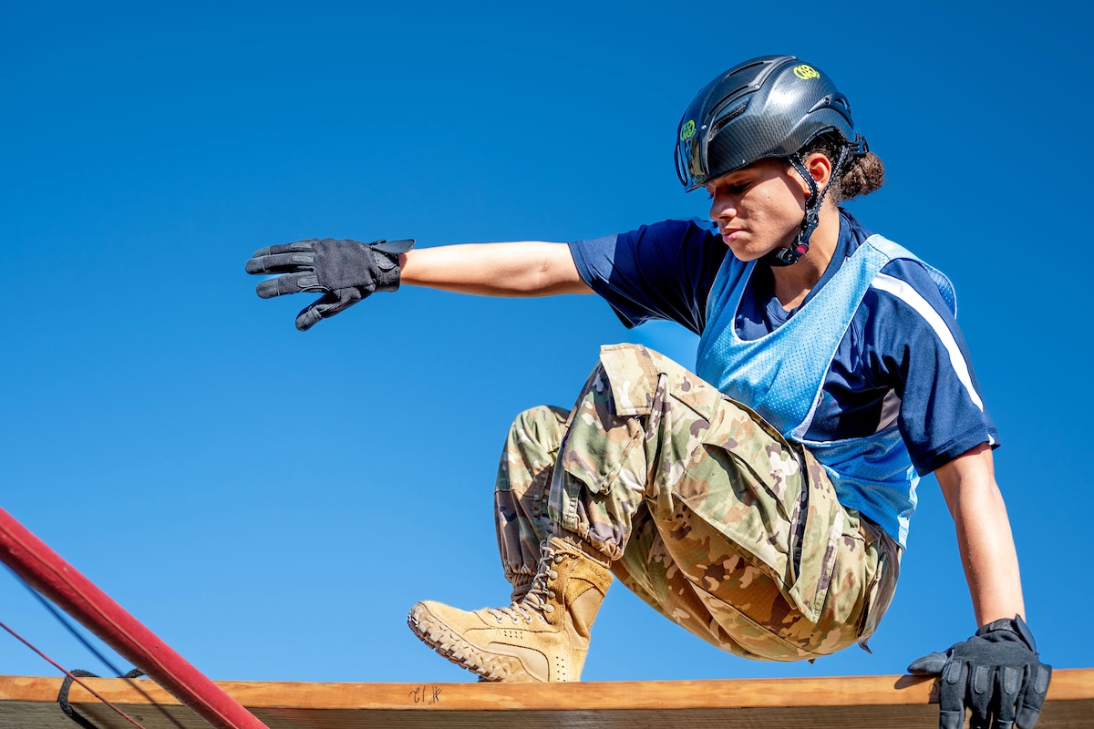 An airman wearing a helmet and gloves climbs an obstacle during daylight.