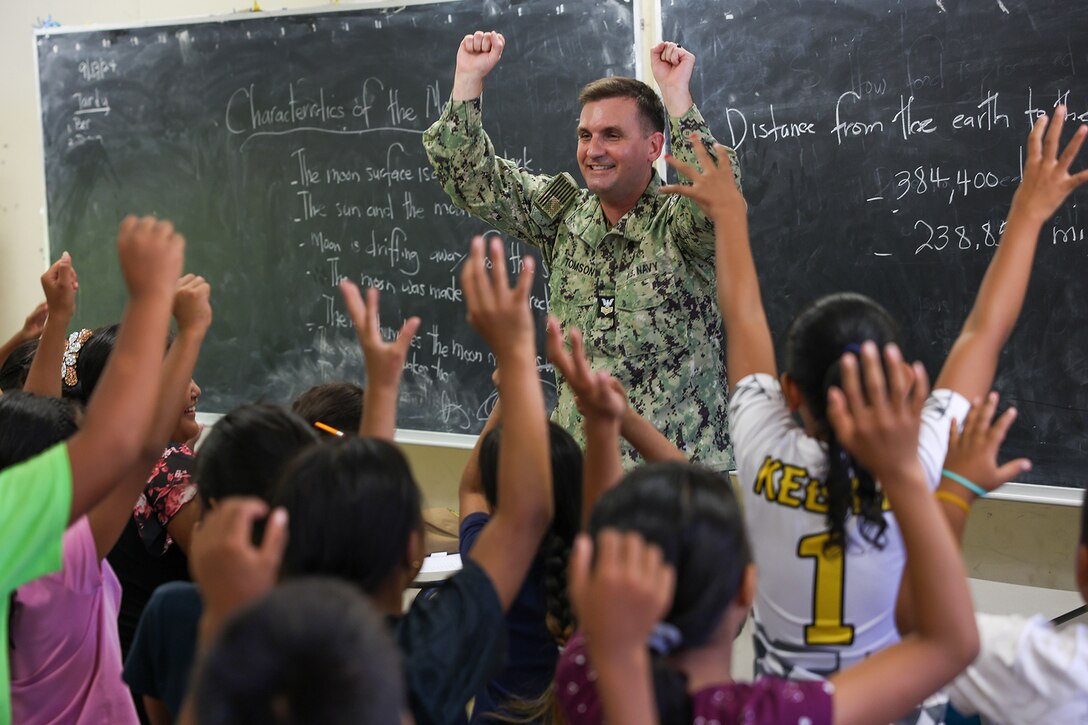 A sailor and a classroom of students raise their hands in unison.