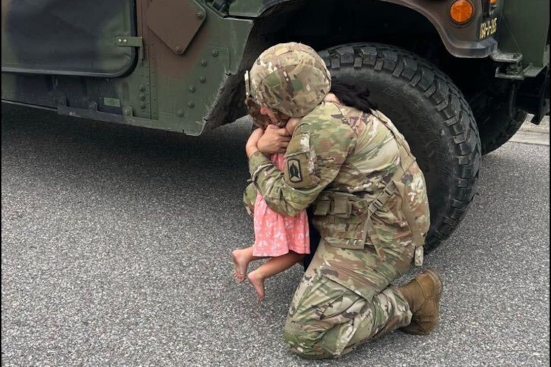 A soldier kneeling on pavement lifts a barefoot child in a pink dress while hugging the child in front of a parked military vehicle.