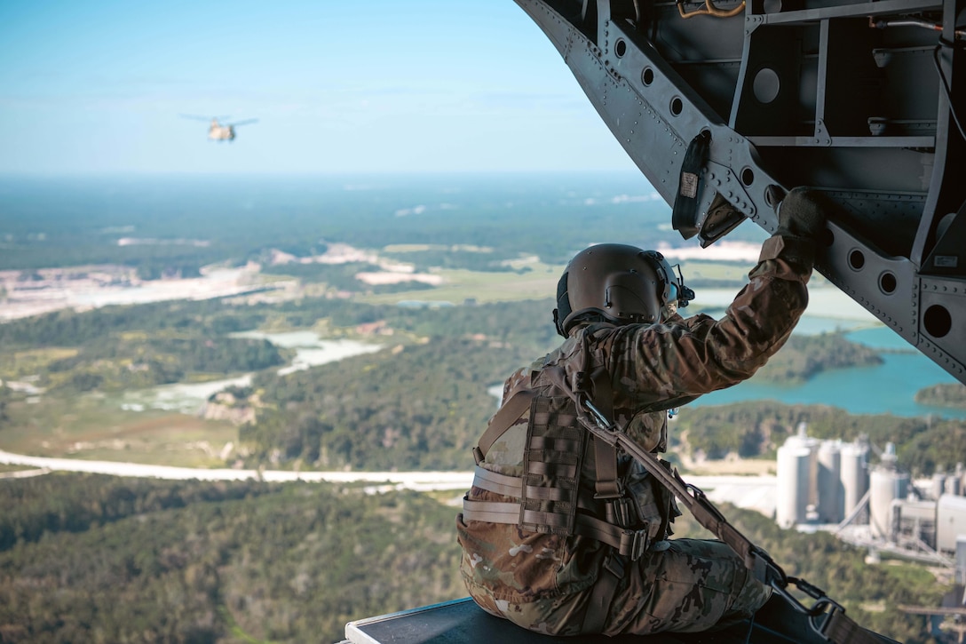 A guardsman sits in the open bay of a Chinook helicopter flying high above buildings. In the distance, another helicopter flies.