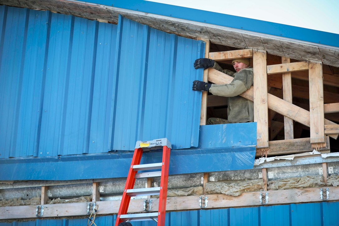 A service member put their arms through an open wooden structure to attach blue shingles during daylight.