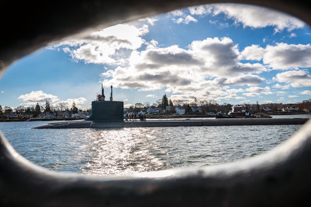 Service members work on top a of submarine as it transits a body of water as seen through the porthole of another ship.