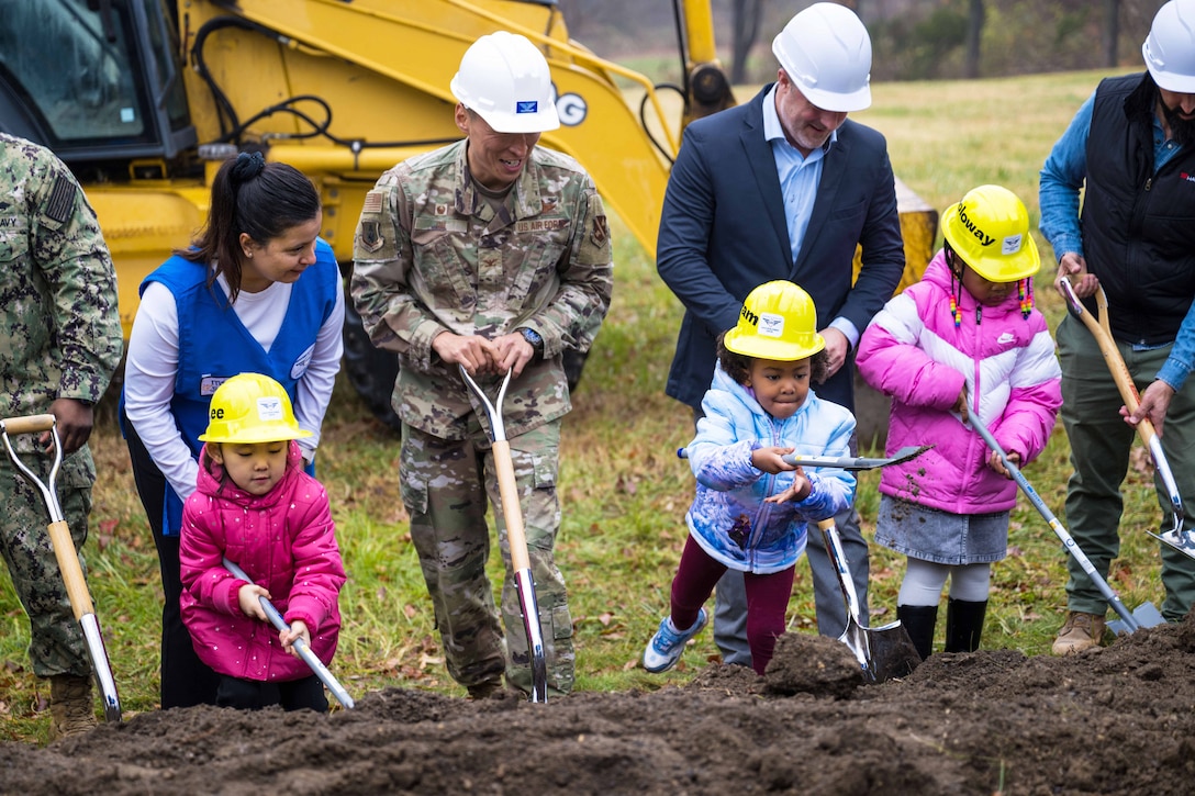 An airman wearing a hardhat shovels dirt with three children and four other adults in a field in front of a construction machine.