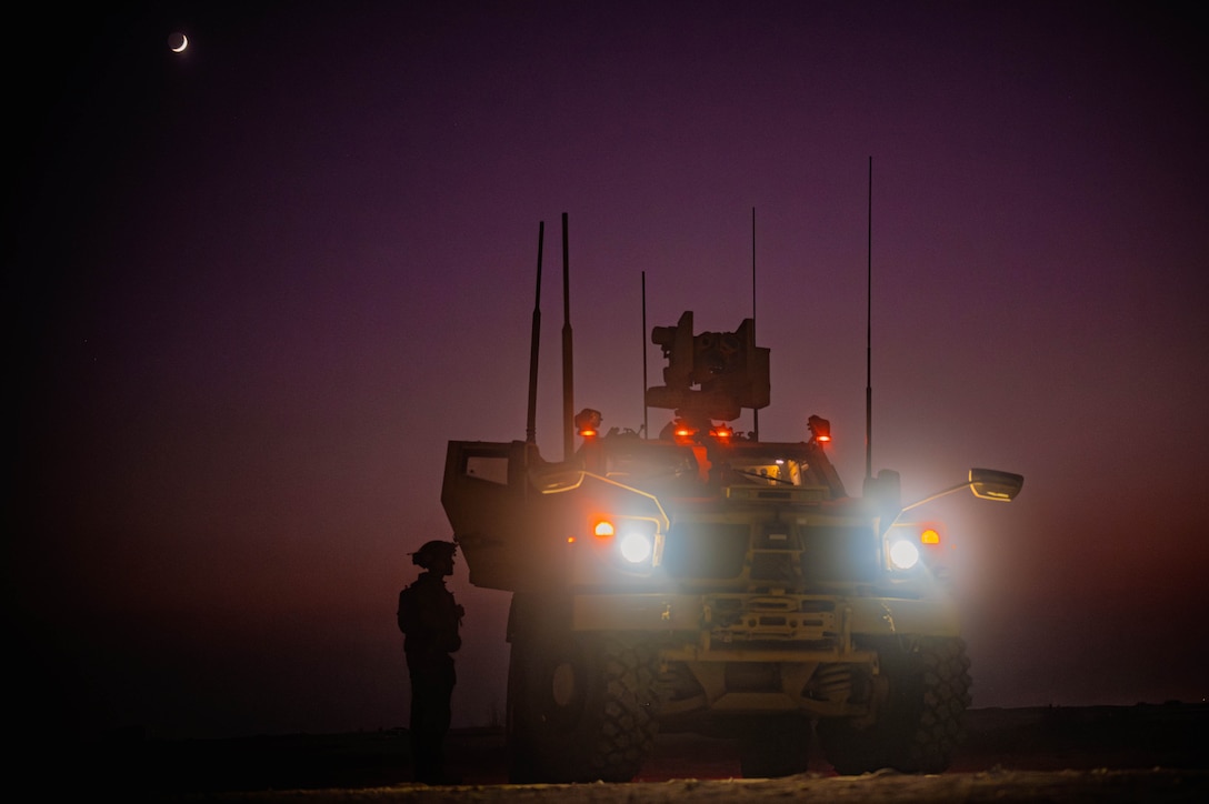 An airman stands next to a military vehicle with headlights on in the dark.