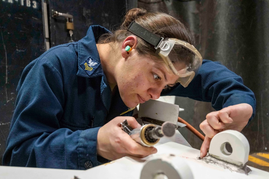 A sailor looks down at a white beam with a crack in it while holding a welding tool.