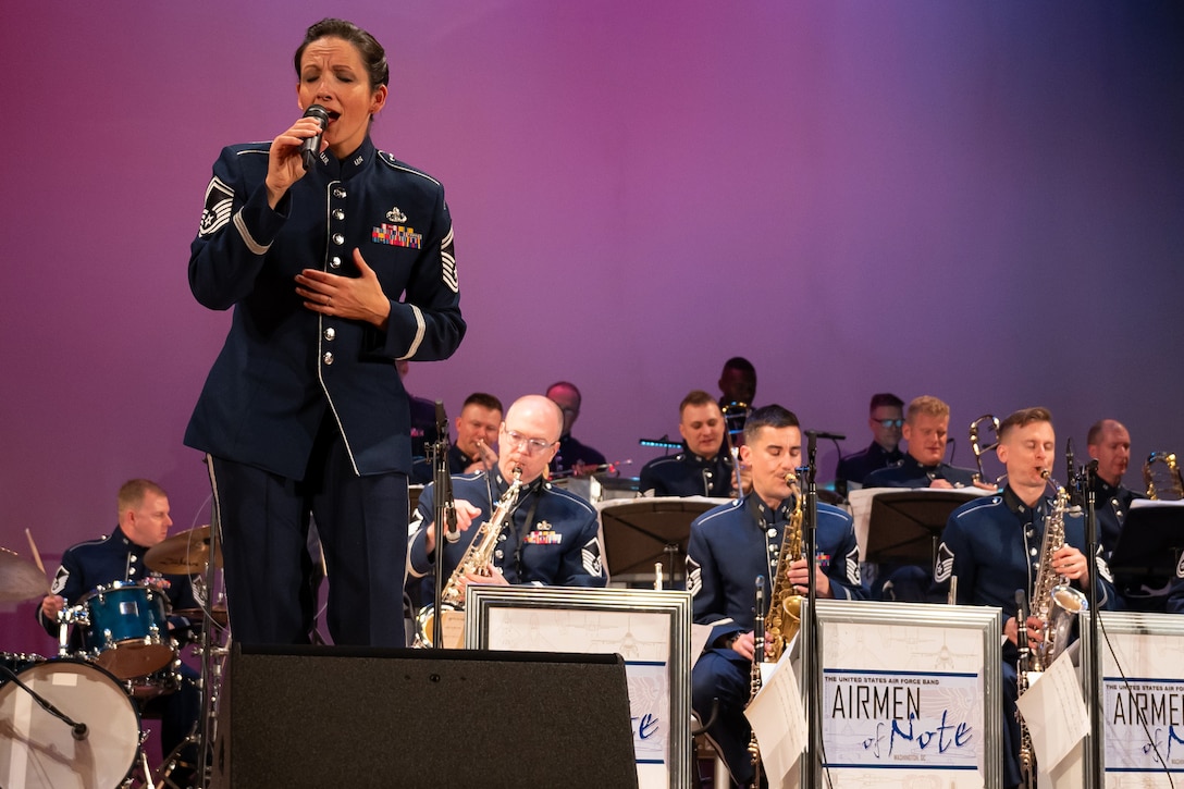 An airman sings on a stage with a microphone while flanked by an ensemble of musicians playing instruments and wearing Air Force attire.