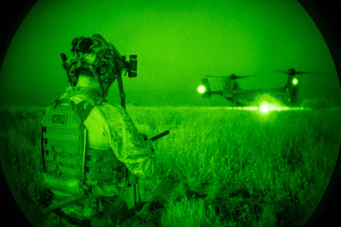 As seen from a night-vision lens, an airman wearing combat gear watches an Osprey aircraft in a field at night.