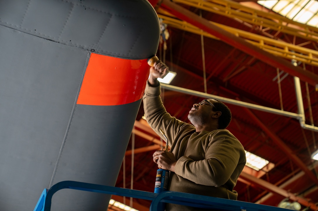 An airman on a raised ladder uses a tool to affix an orange decal to an aircraft.