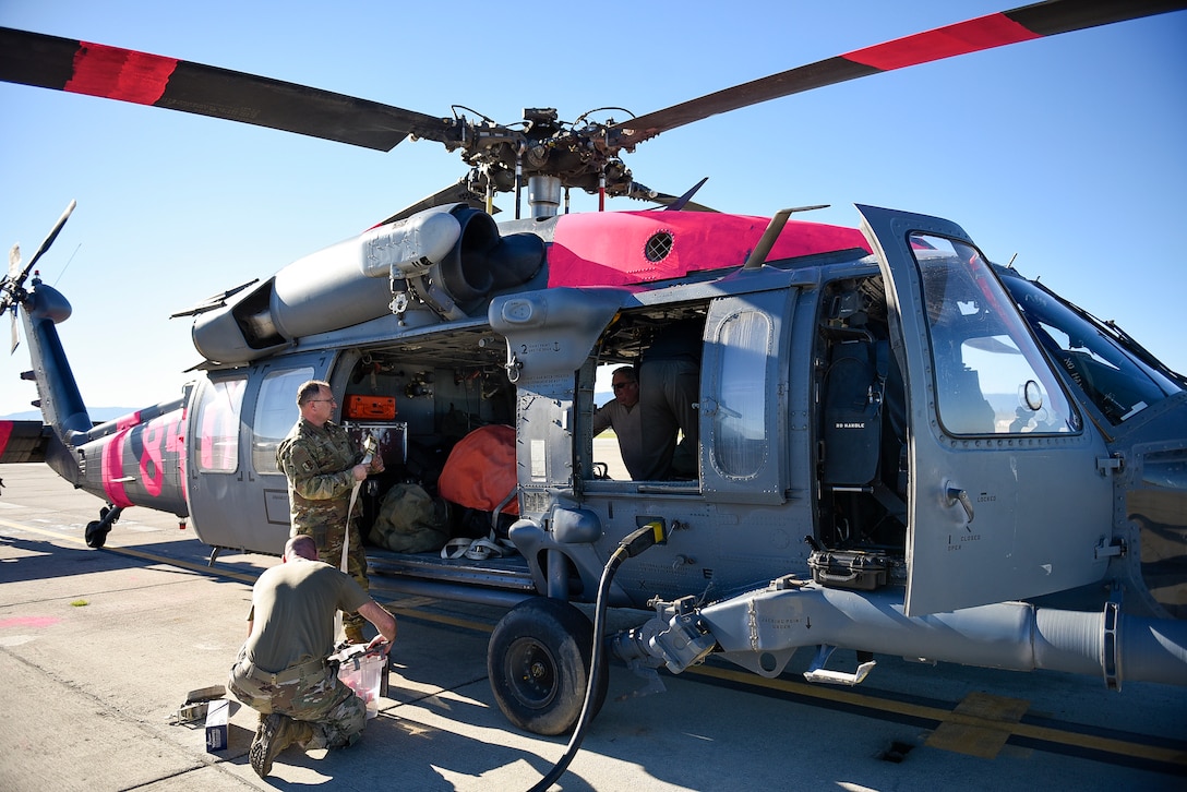 One airman kneels outside a helicopter while two others work on equipment inside.