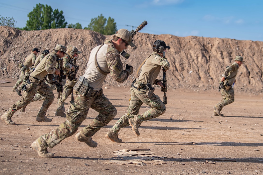 Airmen run through a desert-like area while carrying weapons on a sunny day.