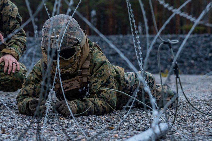 Two Marines on gravelled ground crouch over a cord behind coils of wire.