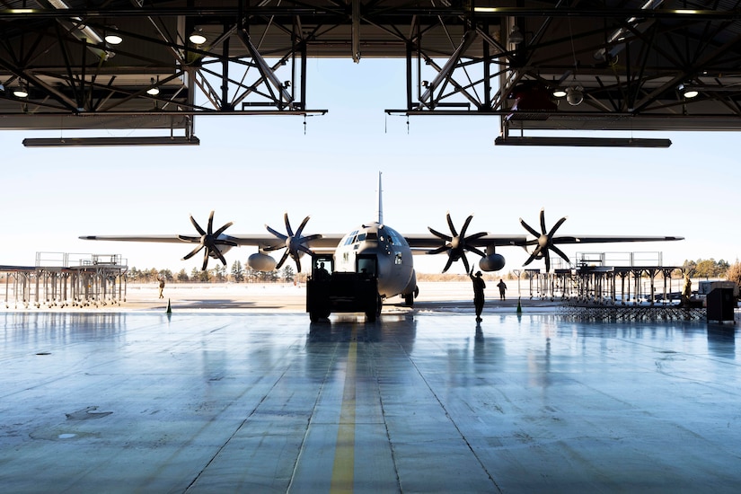 Airmen stand near a military aircraft outside of a hangar on a clear day.