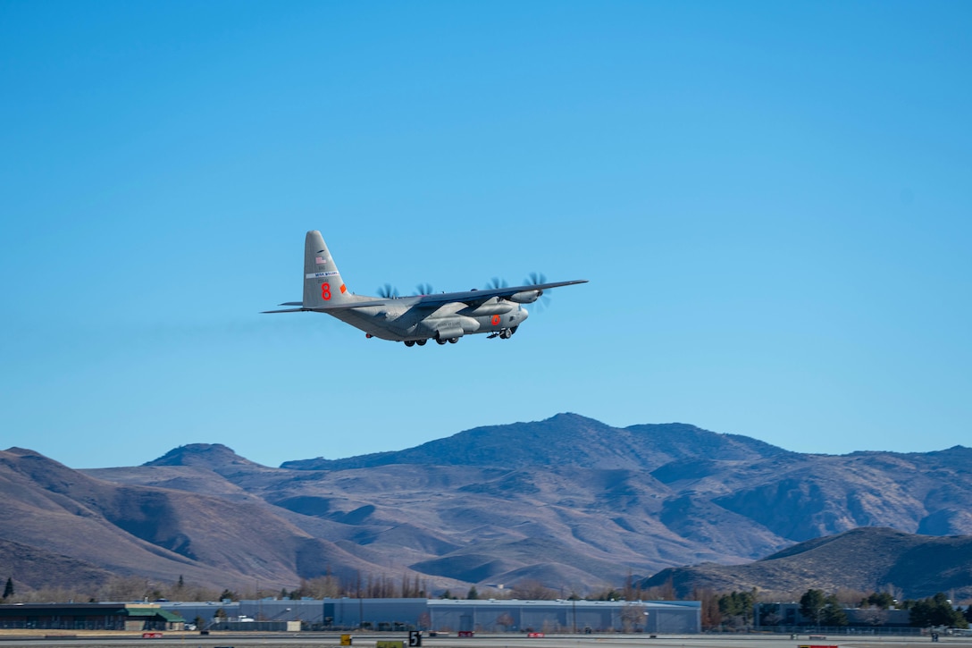 A military aircraft flies above a base against blue skies with mountains in the background.