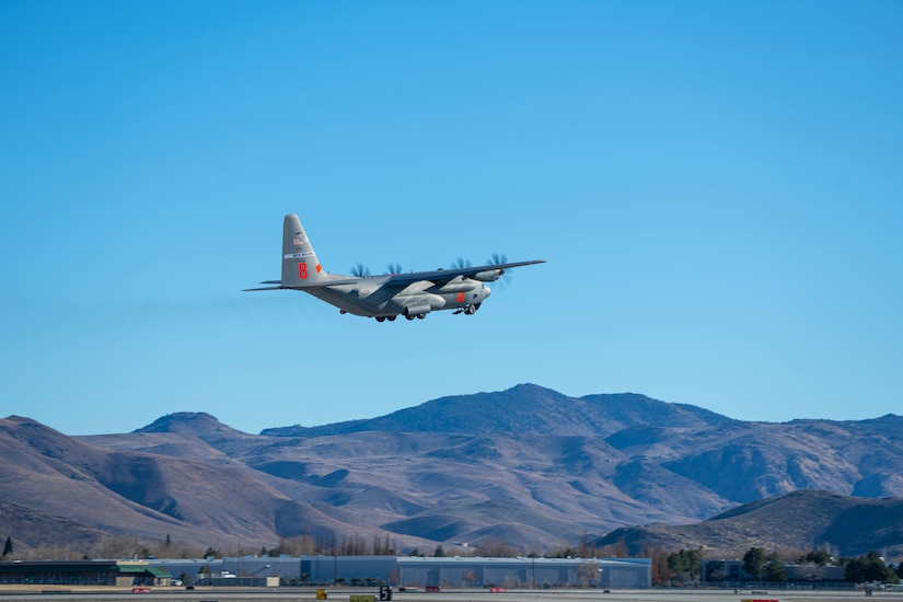 A military aircraft flies above a base against blue skies with mountains in the background.