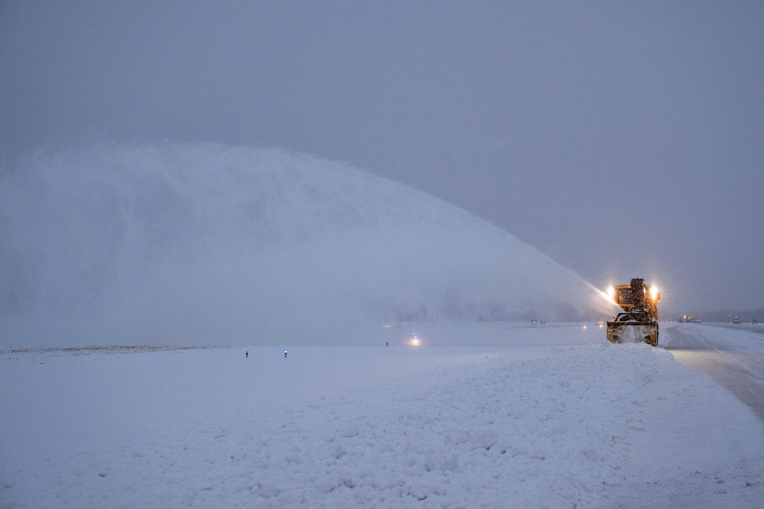 Snow flies in the air as an airman operates a snow blower on a flight line illuminated by yellowish spotlights.