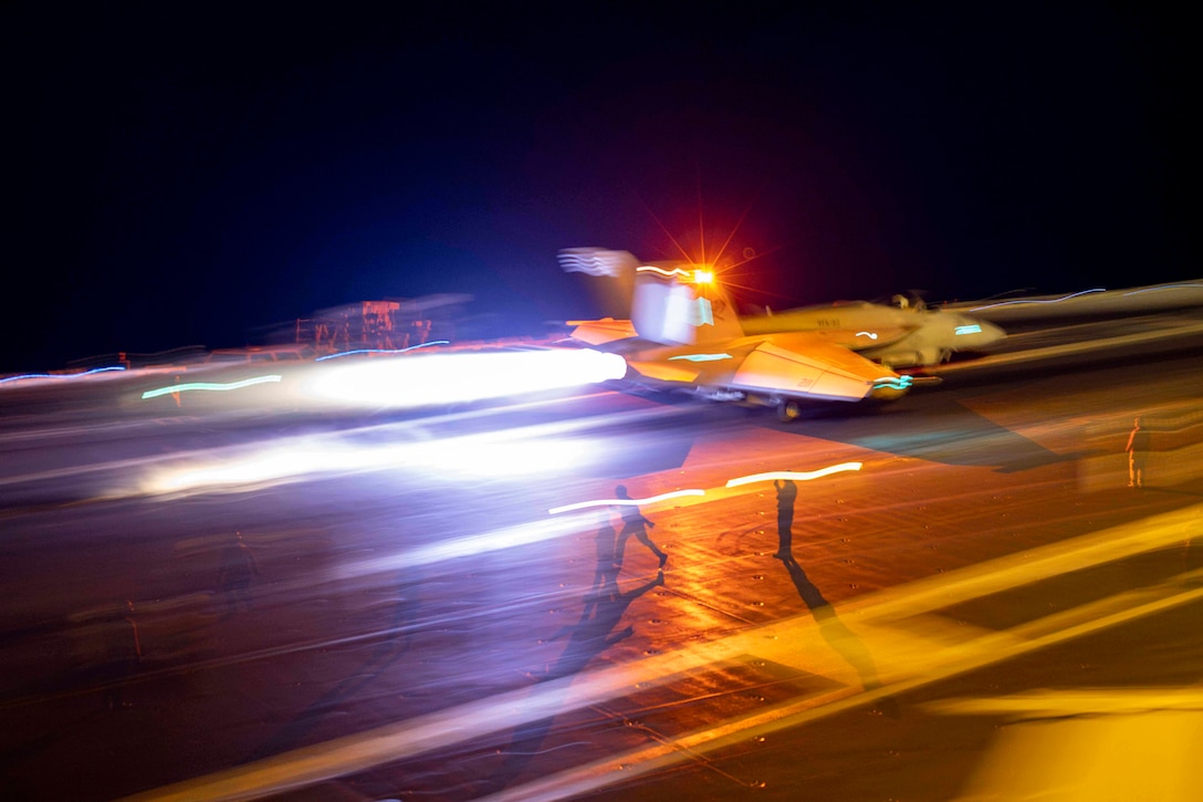 Three sailors signal from the deck as an aircraft takes off from the deck of an aircraft carrier at night leaving streaks of light in its wake.