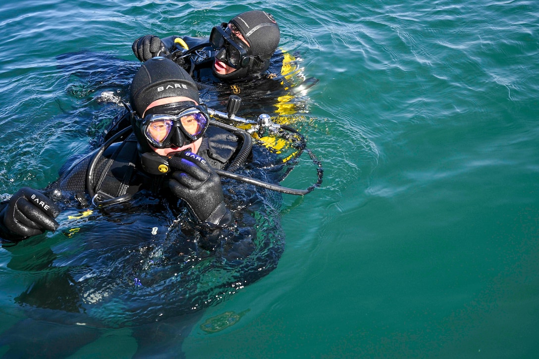 Two sailors in scuba gear float on the surface of ocean water during a sunny day.