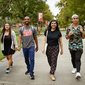 four smiling students walk towards the camera along a path in a university quad