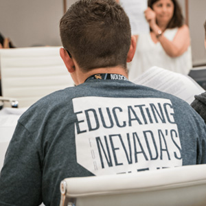 A student wearing a t-shirt that reads "Educating Nevada" listens intently to a lecturer a few feet away