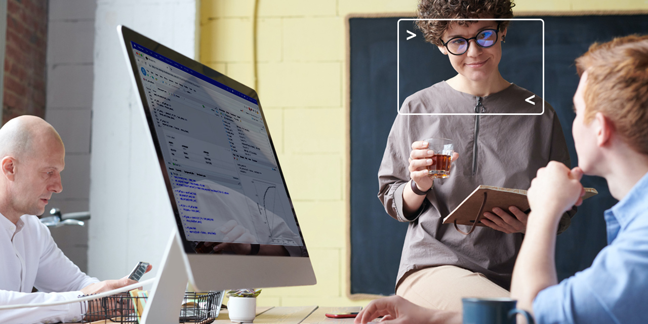 3 people sit a a desk, 2 people are talking near a desktop computer. One of the people has a rectangular computer terminal over her face.