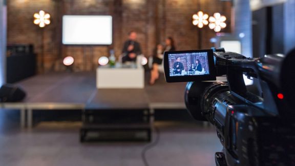 News cameraperson films a group of people in discussion around a coffee table