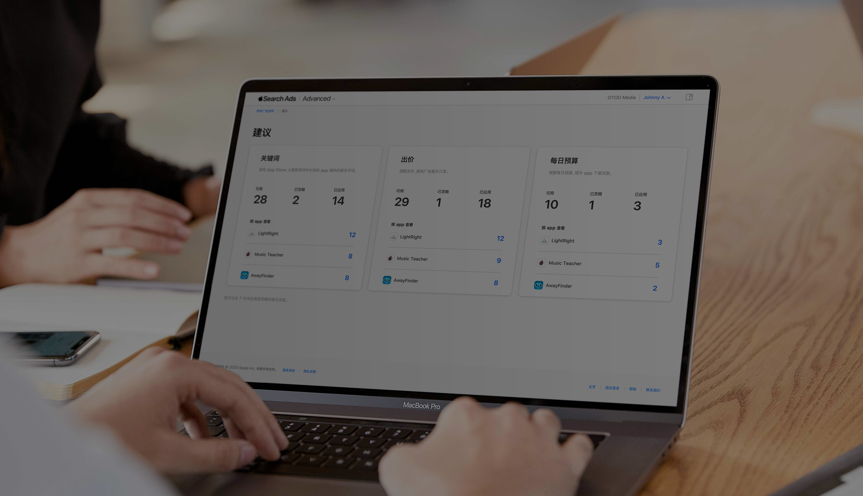 两只手在 MacBook 键盘上打字的特写。屏幕上打开了 Apple Search Ads“建议”页面，并显示按关键词、出价和每日预算划分的推荐。