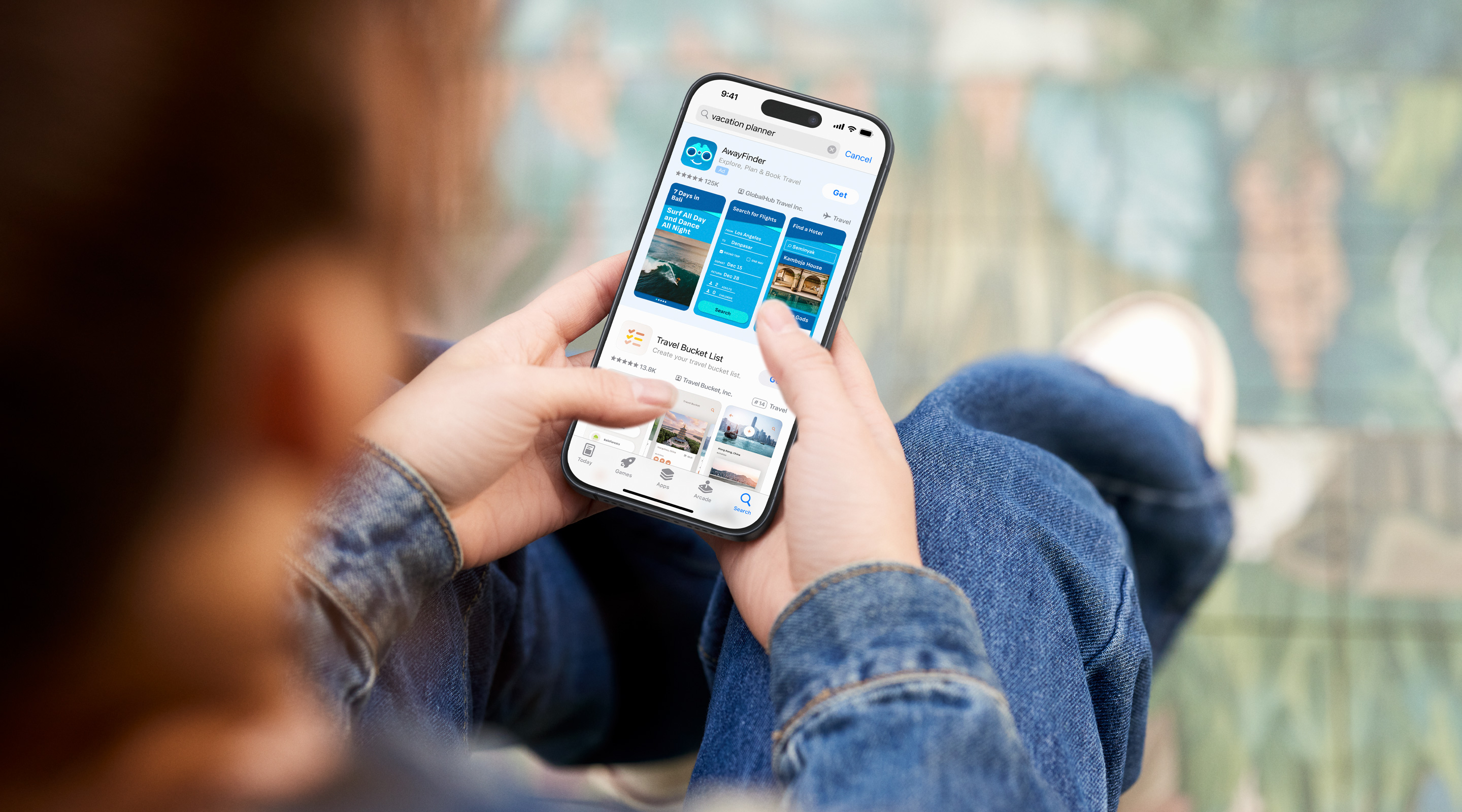 Two hands holding an iPhone. The App Store is open to a search results ad for the example app, AwayFinder, with the term "vacation planner" entered in the search box.